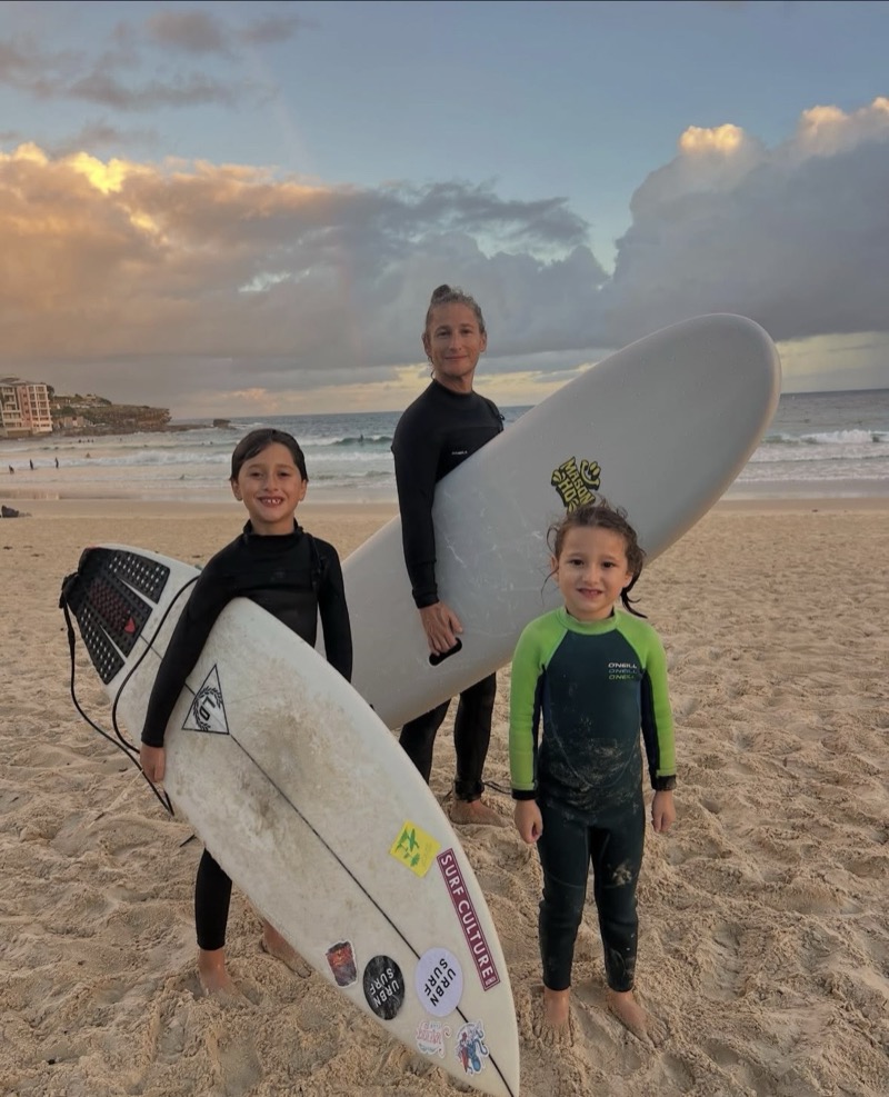 A parent and two kids in wetsuits holding surfboards on the beach at sunset
